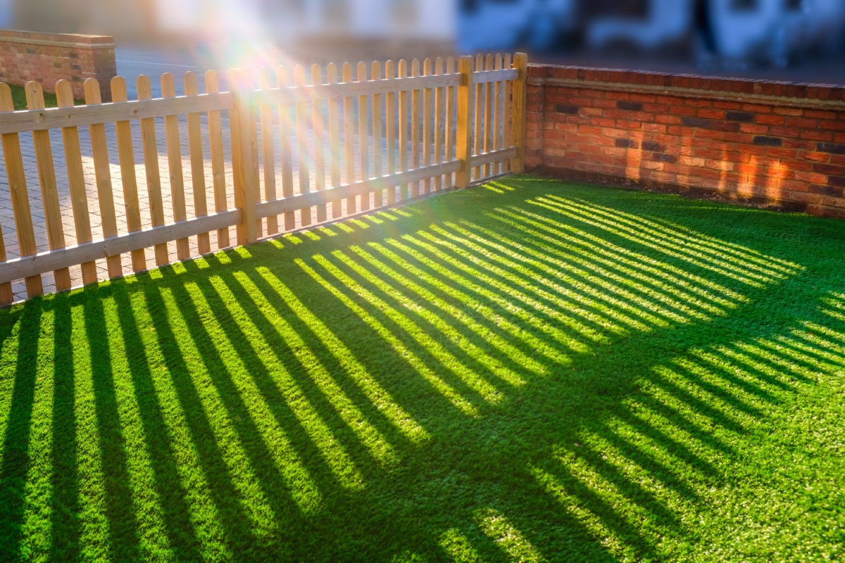 sunshine creating lens flare through a wooden picket fence in a front yard, front garden with artifical grass as a lawn and a red brick perimeter wall.